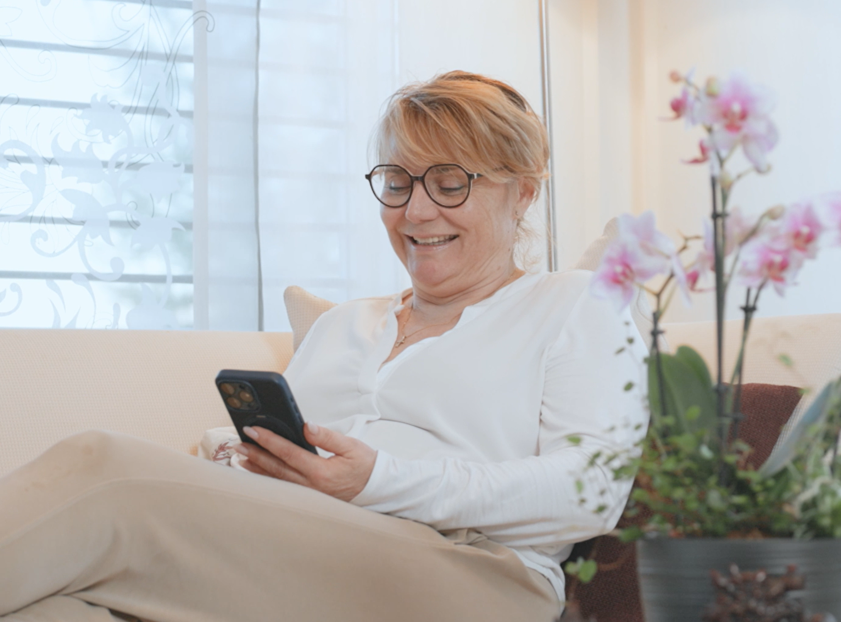 Frau mit Brille sitzt auf dem Sofa und schaut lächelnd in ihr Smartphone